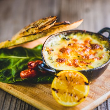 Photo of a small round cast iron skillet of cheese dip served on a small wooden cutting board garnished with a grilled lemon, Toasted French bread slices, green leaf lettuce and sun dried tomatoes 