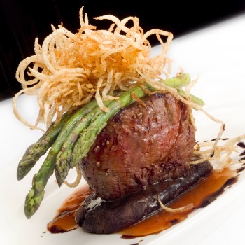 Photo of a dinner entrée with a portabella mushroom, under a filet mignon with asparagus draped over the top and topped with fried onion strings 